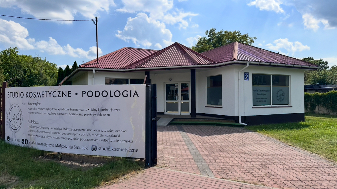 Budynek gabinetu Studio Kosmetyczne Małgorzata Śmiałek przy ul. Żytniej 2 w Łasku · biały dom z bordowym dachem, banner z pełnym menu usług kosmetologicznych i podologicznych obok wejścia.